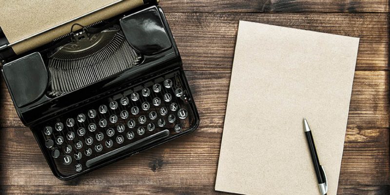 A manual typewriter and. blank piece of paper with a pen, all sitting on a wooden tabletop.